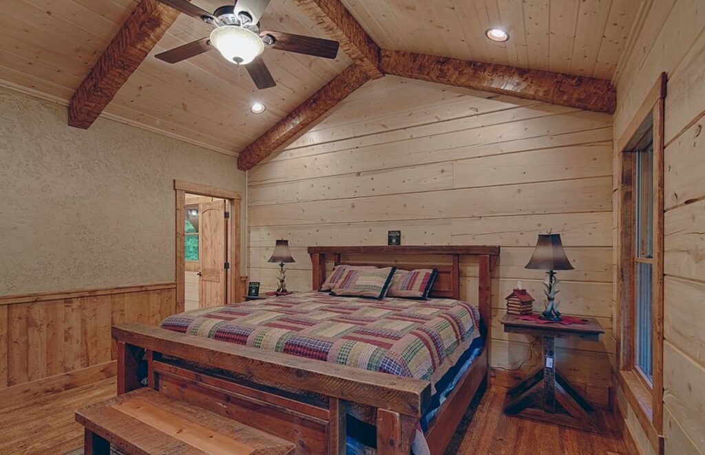 The image showcases a cozy bedroom within a log cabin, complete with rustic charm and cooling feature of a ceiling fan.