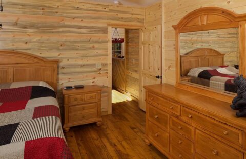 The image showcases a cozy bedroom in one of our log homes, featuring a comfortable bed, a wooden dresser, and an elegant mirror.