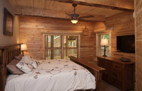 The image displays a cozy, rustic bedroom within one of our log homes, featuring a comfortable bed and an overhead ceiling fan.