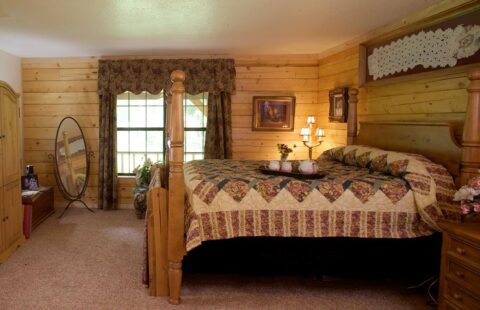 The image displays a cozy, rustic-style bedroom inside a log home featuring a comfortable bed and matching wooden dresser.