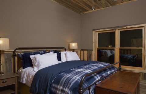 The image showcases a cozy bedroom in one of our log homes, beautifully adorned with wood paneling and an inviting bed.