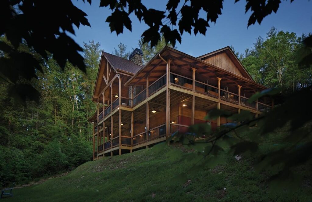The image captures a beautifully crafted log cabin nestled into the tranquil woods, glowing warmly as dusk falls.