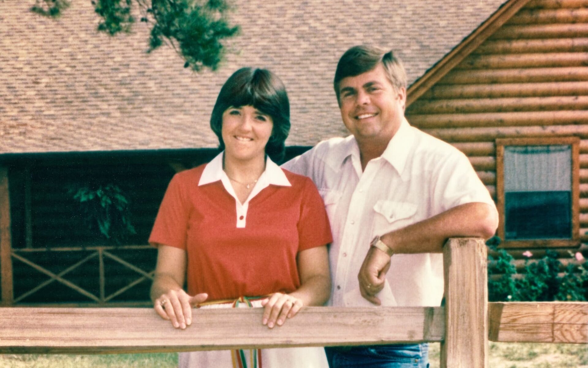 A man and woman are standing beside a sturdy fence, with our beautifully constructed log home showcased in the background.