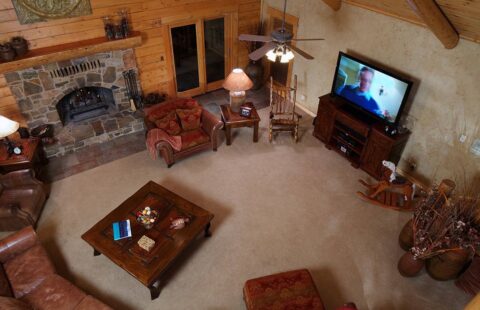 The image showcases a cozy, rustic living room in one of our log homes featuring a roaring fireplace and modern TV unit.