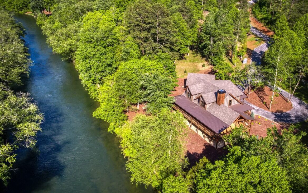 The image showcases a stunning bird's-eye view of one of our beautifully crafted log homes nestled beside a serene river.
